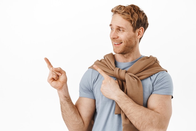 Smiling redhead man with muscles big biceps, pointing at upper left corner, showing advertisement on product on copy space, standing over white wall