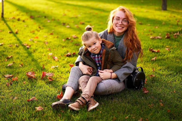 Smiling redhead woman and little boy sitting on grass lawn in city park on warm autumn day young mom