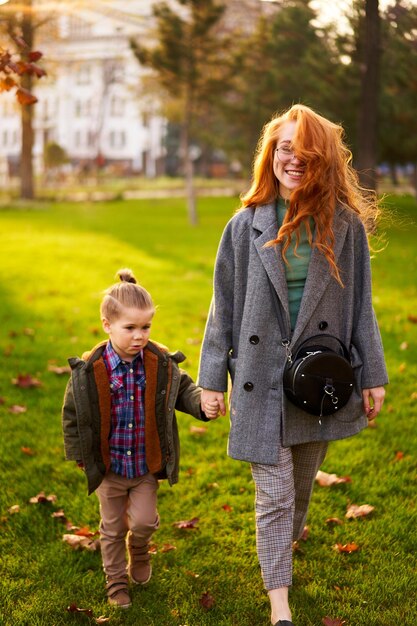 Smiling redhead woman and little boy walking on green grass lawn in city park on warm autumn day Young mom goes with her son they have fun and hold hands on sunny fall day Foliage on green lawn