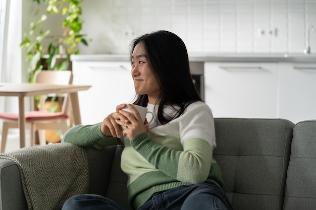 Smiling relaxed female sitting on sofa at home holding cup of tea and relaxing after stressful day