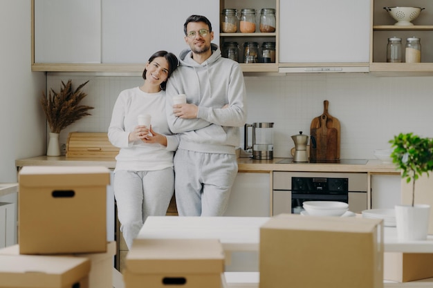 Smiling relaxed husband and wife drink coffee at kitchen hold paper cups with hot beverage dressed in casual outfit relocate in new apartment pile of cardboard boxes with property in foreground