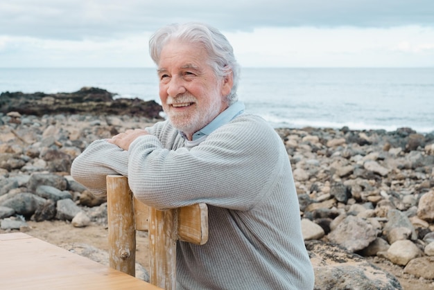 Smiling relaxed mature senior man with beard sitting in outdoors at sea White haired caucasian elderly grandfather sitting at the beach on a wooden chair with crossed arms