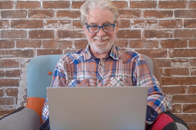 Smiling relaxed senior man sitting at home on armchair using laptop computer. Brick wall in background