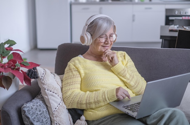 smiling relaxed senior woman wearing headphones sitting on sofa at home using laptop