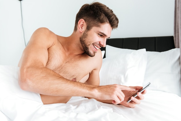 Smiling relaxed young man lying and using mobile phone in bed