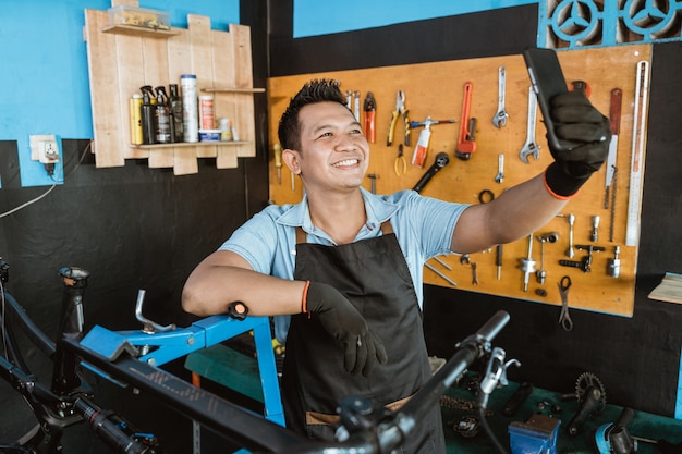 A smiling repairman in an apron selfie using a cellphone while repairing a bicycle