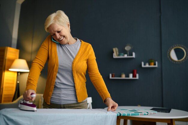 Smiling retired aged woman ironing clothes on board. Happy retirement, domestic work