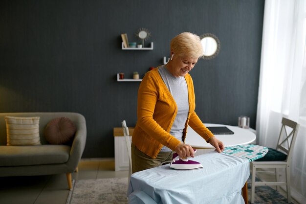 Smiling retired aged woman ironing clothes on board. Happy retirement, domestic work