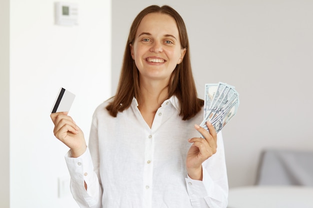 Smiling rich woman wearing white casual style shirt, winning lottery, holding credit card and dollar banknotes in hands, looking at camera with happy expression, posing in light room at home.