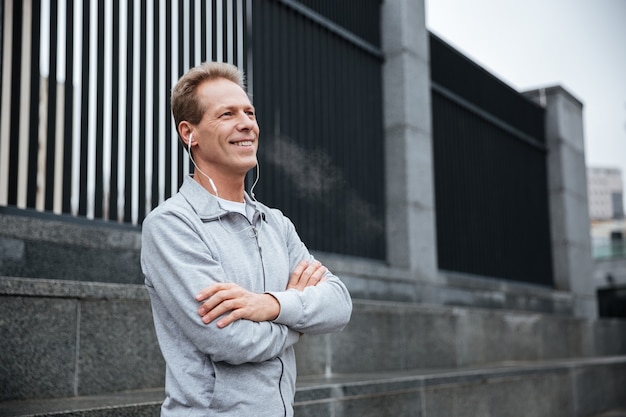 Smiling Runner in gray sportswear and headphone standing with arms crossed on the street.