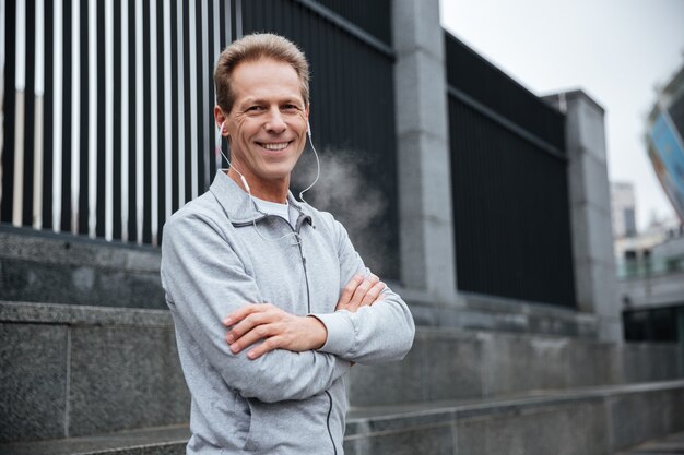 Smiling Runner in gray sportswear and headphone standing with arms crossed on the street