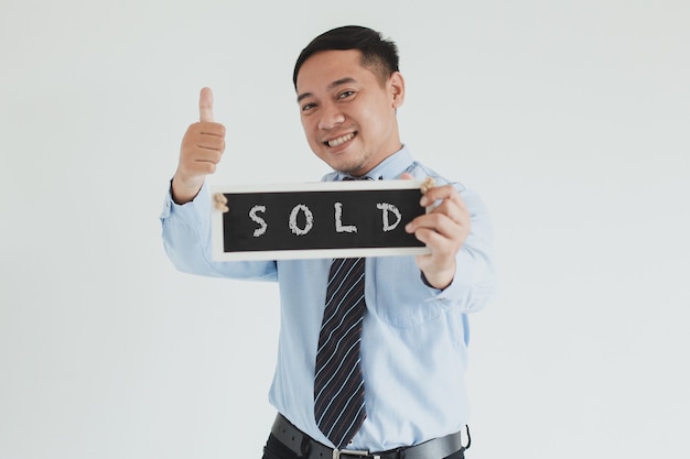 Smiling sales man wearing blue shirt and tie posing with thumb up and showing SOLD sign board at camera on white background