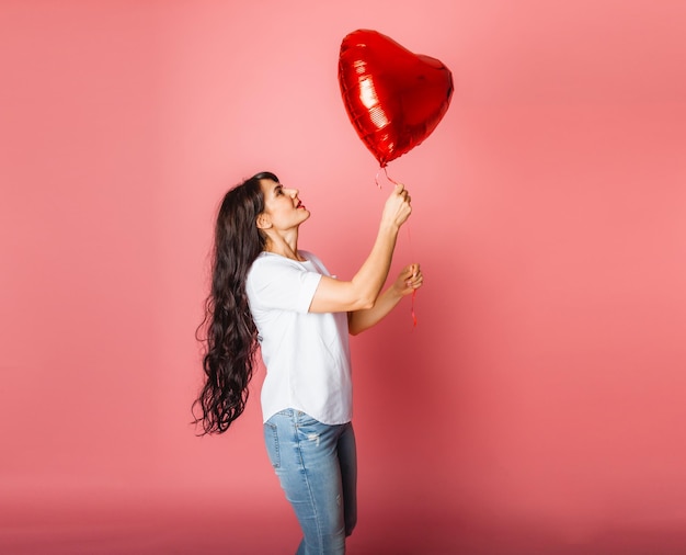 Sorridente ragazza caucasica soddisfatta in posa con un palloncino rosso a forma di cuore su sfondo rosa. san valentino