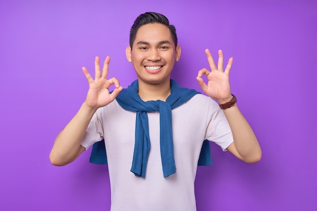Smiling satisfied and cool young Asian man 20s wearing white tshirt casual clothes showing okay ok gesture isolated on purple background People lifestyle concept