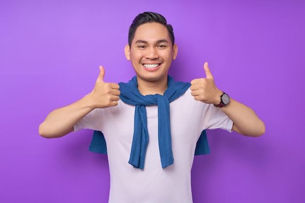 Smiling satisfied and cool young asian man 20s wearing white tshirt casual clothes showing thumbs up like gesture isolated on purple background people lifestyle concept