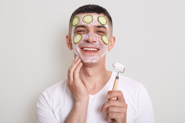 Smiling satisfied man wearing white t shirt posing with
moisturizing mask and slices of cucumber on his face holding
massage rolling for cosmetology procedures isolated over gray
background