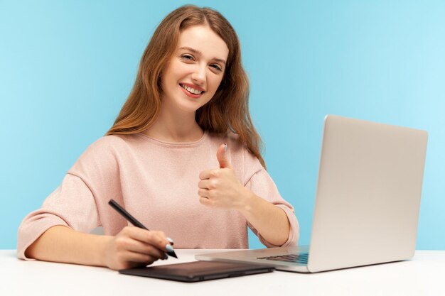 Smiling satisfied woman creative designer sitting at workplace with graphic tablet and laptop showing thumbs up while working on professional digital art equipment indoor studio shot isolated