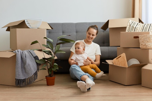 Smiling satisfied woman wearing white t shirt and jeans sitting on floor near cough with her toddler daughter relocating to a new apartment unpacking personal belongings