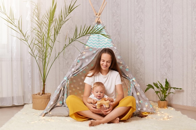 Sorridente donna soddisfatta con lunghi capelli scuri che indossa una maglietta bianca seduta in tenda peetee con il suo bambino e tiene in mano un cavallo di legno che gioca con la figlia del bambino a casa a wigwam