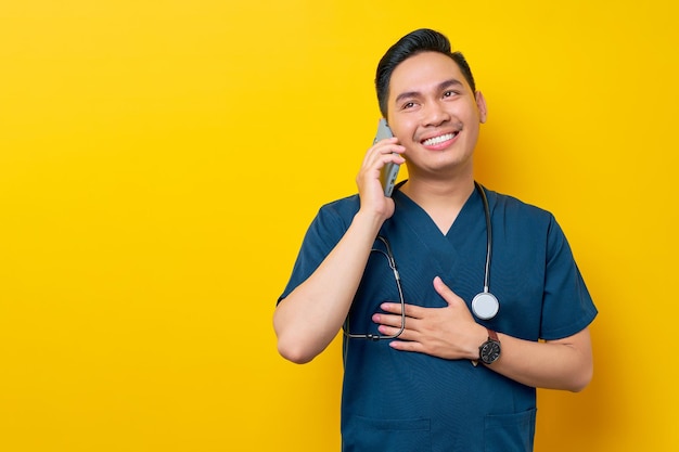 Smiling satisfied young Asian male doctor or nurse wearing blue uniform talking on mobile phone with patient isolated on yellow background Healthcare medicine concept