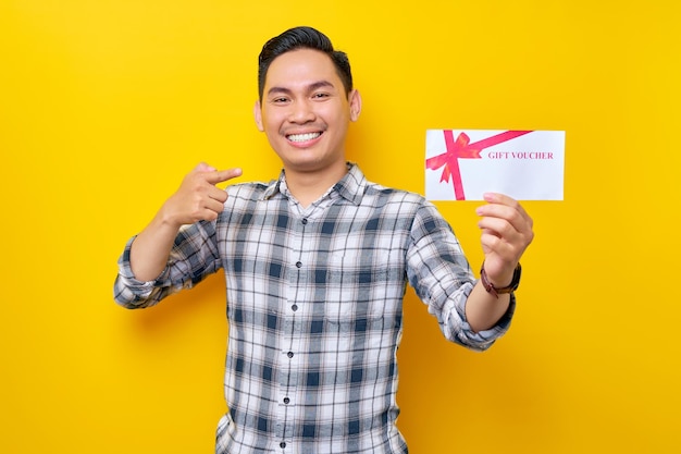 Smiling satisfied young Asian man wearing a white checkered shirt pointing a finger at gift voucher isolated on yellow background People Lifestyle Concept