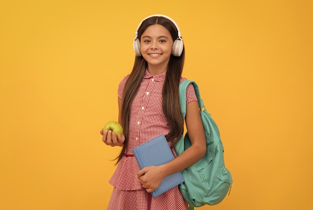 ヘッドフォンで笑顔の小学生は、ランチデトックスのためのアップルとバックパックとワークブックを運ぶ