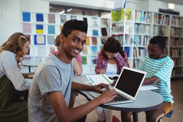 Smiling schoolboy using laptop with his classmates studying