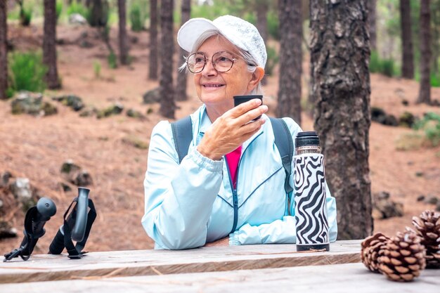 숲 속 트레킹을 하며 나무 테이블에 앉아 물을 마시는 웃고 있는 활동적인 노인 여성