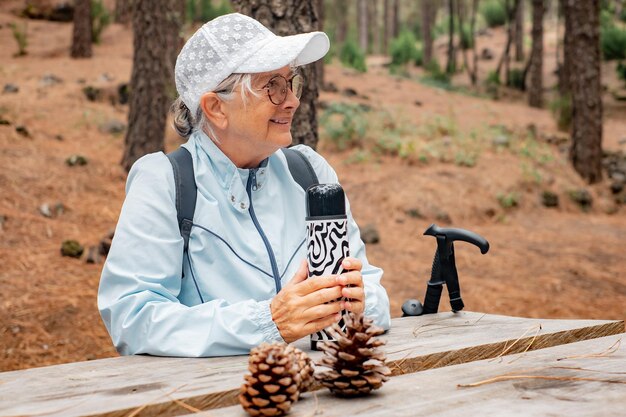 Smiling senior active woman in trekking day in the woods sitting at a wooden table to rest and drink