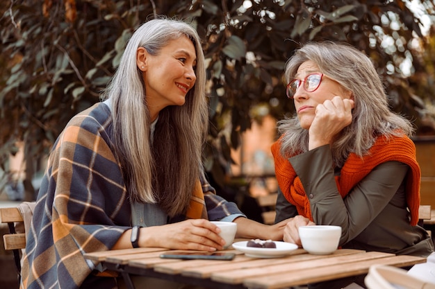 Smiling senior asian woman holds hand of upset friend sitting together in street cafe