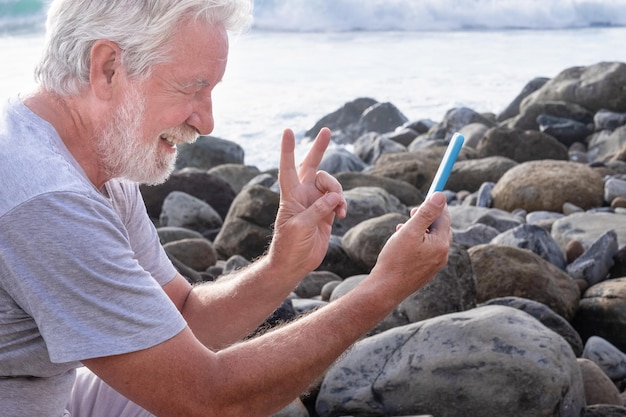 海に面した岩のビーチに座って携帯電話でビデオ会話をしている笑顔のシニアひげを生やした男