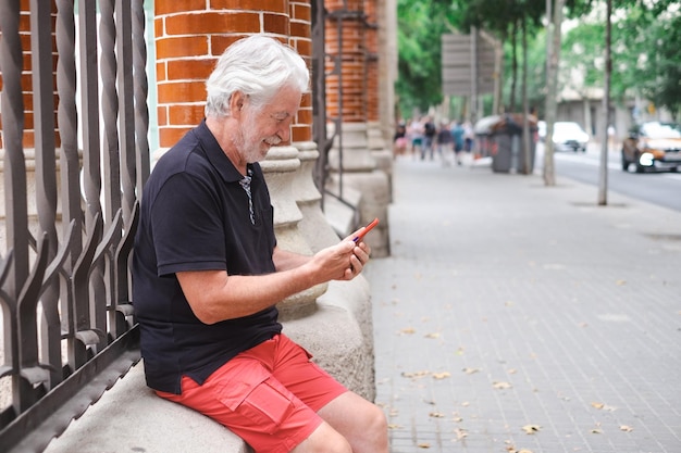 Smiling senior bearded man sitting outdoor historic building in Barcelona using mobile phone consulting next destination Freedom vacation concept