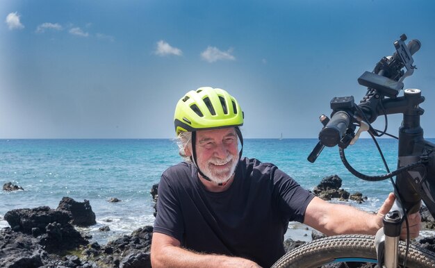 Smiling senior bearded man with helmet and electric bicycle in outdoors at sea Caucasian happy grandfather enjoying retirement and freedom checks his bike before healthy ride