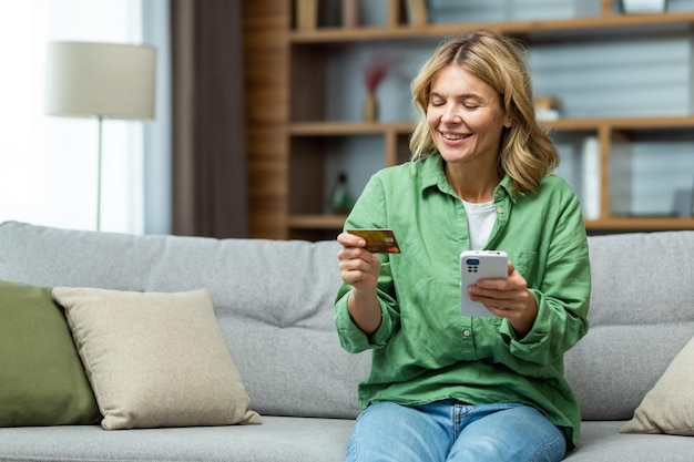 Smiling senior beautiful woman sitting on sofa at home holding a phone using a credit card smiling