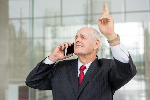 smiling senior business man looking away, calling on smartphone and raising hand