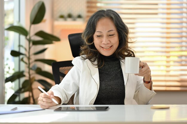 디지털 태블릿으로 집에서 온라인으로 일하고 뜨거운 커피를 마시는 웃고 있는 고위 여성 사업가