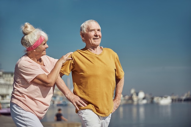 Smiling senior Caucasian lady with a headband supporting her husband performing a warm-up exercise outdoors