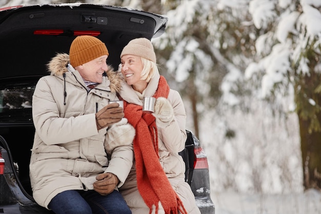 Улыбающаяся пожилая пара наслаждается чашкой горячего кокоса на открытом воздухе зимой, сидя в багажнике машины