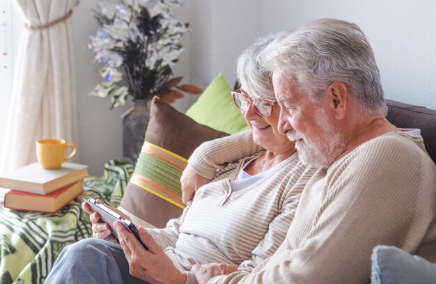 Smiling senior couple hugging sitting on sofa at home looking together at digital tablet browsing on internet Lifestyles retirement concept