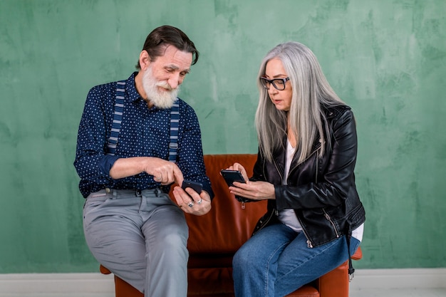 Smiling senior couple, stylish bearded man and pretty gray haired lady, enjoying review of photos on their smartphones