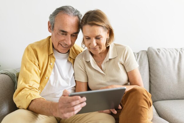 Smiling senior couple using tablet computer surfing internet watching movie online together sitting