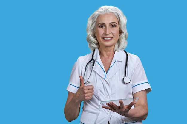 Smiling senior doctor using transparent digital tablet and showing thumb up gesture on blue backgrou