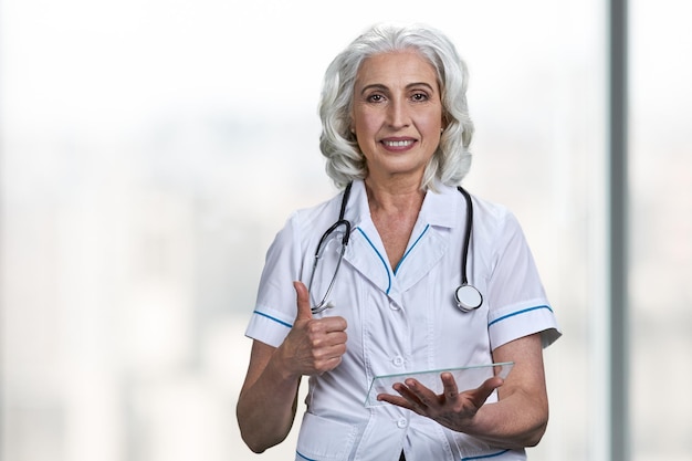 Smiling senior doctor using transparent digital tablet and showing thumb up gesture. People, modern technology and future innovation concept.