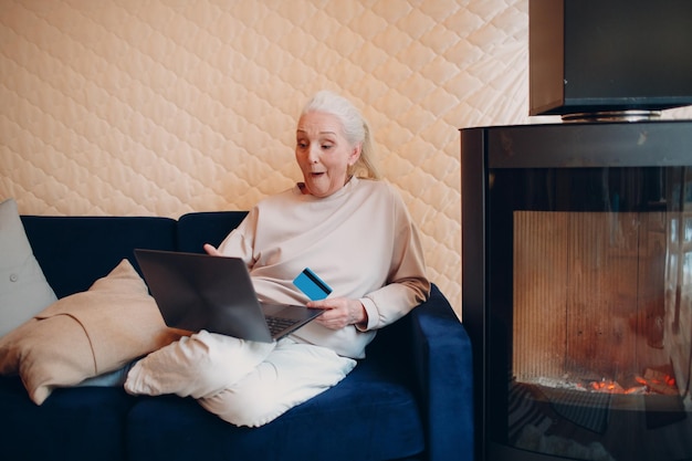 Smiling senior ederly woman using credit bank card and laptop on sofa near fireplace Online shopping concept