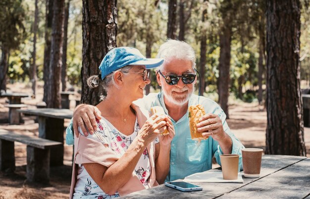 Smiling senior family couple enjoying a sandwich sitting at a wooden table in the woods