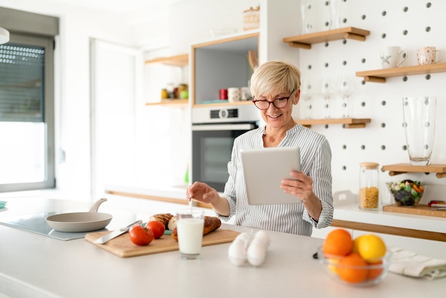 Smiling senior gray hair woman with eyeglasses looking at digital tablet while making healthy breakfast in kitchen