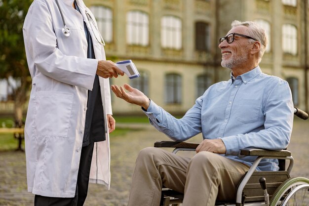 Smiling senior handicapped man reaching out hand for doctor to check fever by digital thermometer