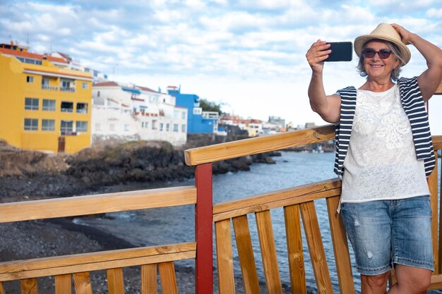 Smiling senior lady with straw hat using phone for a selfie\
with horizon over the harbour in background relaxed people in sea\
excursion cheerful retirement concept