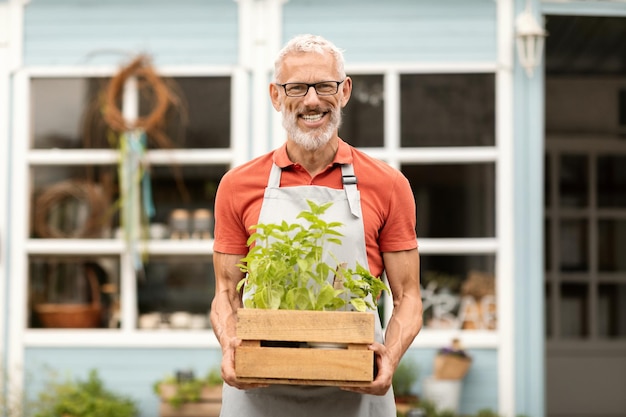 Photo smiling senior male gardener holding crate with plants while walking outdoors