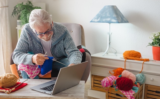 Uomo anziano sorridente che si diverte a lavorare a maglia a casa seguendo il tutorial di maglia online per rilassarsi e godersi l'attività di svago seduti su una poltrona di sesso maschile che lavora a maglia con l'aiuto di classe del computer